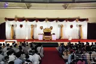 A large indoor hall set for a gathering, with a raised red-carpeted stage backed by white draped fabric, maroon swags and orange-and-red floral garlands. Four tall gold-painted pillars flank the stage, and an ornate gold throne with a dark cushioned seat stands centrally, with two lilac-draped side tables holding flowers. Rows of seated devotees fill the foreground on a red carpet and mats; many wear white headbands or head cloths and white or light clothing, with some in darker shirts. The photo is a scene with several dozen people visible, seen from behind them facing the stage.