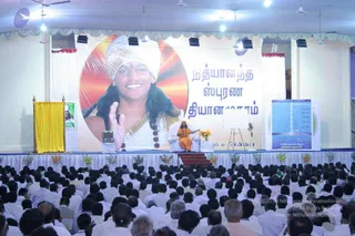 A large audience of many seated devotees, mostly men in white clothing, fills a hall facing a stage. On the stage, a figure in orange robes sits on a white chair behind a microphone, with a yellow curtained podium at the left. Behind him, a large banner shows a smiling man in a white turban and gold-bordered white garment with a rudraksha mala, beside Tamil text. A tall display panel at right reads Nithyananda State and Ananda Gandha. Flower arrangements line the stage edge and ceiling fans and lights hang overhead.