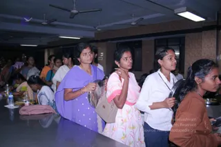 Photo of women and young girls queuing along a long steel serving counter in an indoor hall with ceiling fans and fluorescent lights, holding metal plates for a meal. A woman in a lavender-purple salwar with a gold necklace and red-orange bangles holds a steel plate at the front. Next, a young woman in a pink and white floral dress with a beige handbag holds a plate, followed by a girl in a white shirt with a thin chain holding a steel plate, and a girl in a rust-brown top at the right edge. Bottles of water sit on the counter, and more people stand in the background. The image appears to be from a person Spurana convention meal line.
