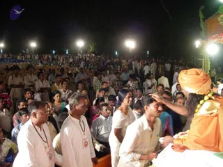 A large night-time outdoor gathering of many devotees standing and seated under bright floodlights on a field. In the right foreground, a figure in an orange turban and orange robe with a long dark hair and a garland of green beads places a hand on the forehead of a young man in white. A man in white with a garland of tulsi-like beads stands at left, and a woman in a white sari with an orange-red necklace stands near the center. Orange canopy and green foliage frame the right edge. Many people fill the background.