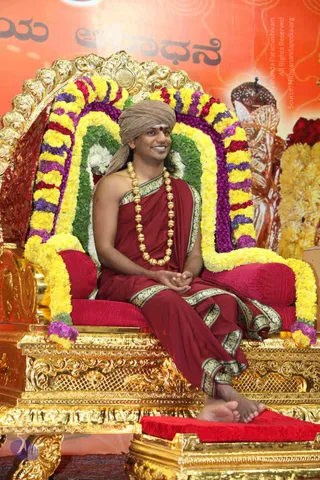 A smiling man in a tan turban with a red bindi and a maroon robe with gold-embroidered border sits on an ornate gold throne (peetha) decorated with garlands of yellow marigolds, purple and red flowers, and green leaves. He wears a long chain of large gold beads around his neck and has bare feet resting on a red cushioned footstool. Behind him a red-orange backdrop shows Kannada script and a painted figure. The setting suggests a formal ceremonial seat.