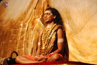 A seated man sits cross-legged in meditation on a red-covered platform in front of a gold-and-cream draped backdrop, eyes closed and hands resting in his lap. He is bare-chested with a leopard-print shawl draped over his shoulder, wears a heavy gold necklace with rudraksha beads and a red-orange dhoti, and has long swept-back hair with a rudraksha crown and white vertical tilak marks. A black kamandalu-style pot sits to his left. Visible cues match the a person appearance profile: long hair, rudraksha adornments, ornate gold necklace and a leopard shawl, and he is physically present in the scene.