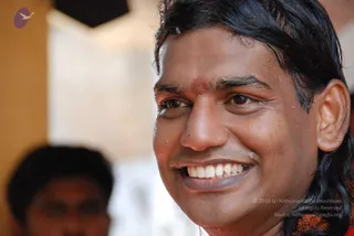 Close-up photograph of a smiling man with long, wet-looking dark hair swept back, a small red-orange tilak mark on his forehead, and water droplets on his face and cheek. A blurred second person stands in the lower left background against a pale wall. The man's shoulder shows a reddish-orange garment.