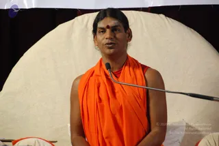 A man seated in front of a large cream-coloured circular backdrop speaks into a gooseneck microphone. He wears a saffron-orange shawl wrapped over one shoulder and draped across the chest, over a red undergarment, and has a small red bindi on his forehead and dark hair pulled back. Cushions are visible at the lower edge of the frame, and a white banner hangs across the top. The setting suggests a seated yoga discourse session. Whether this is the a person named in the page cannot be confirmed from appearance alone, so he is described by role as a speaker.