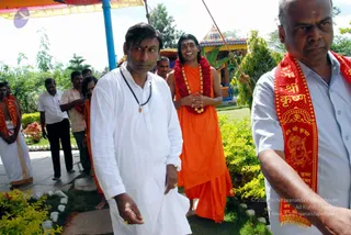 Outdoor photo of a garden path at a temple complex under a yellow-and-red canopy. In the centre, a man in a white dhoti and shirt with a dark rudraksha-style mala walks ahead. Behind him, a smiling figure in saffron-orange robes wears a red flower garland and gold beaded necklace, hands joined at the chest, with bare feet on the grass; the appearance is ambiguous and the role is best described as a sannyasi. To the right, a man in a white shirt wears a red shawl printed with Sanskrit text and a deity image. Background shows a colourful temple gopuram, green shrubs and palm trees, with several men in white and saffron behind them.