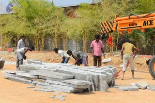 A construction site in daylight with a large pile of grey-white cut granite slabs on bare orange-brown earth. Five workers handle the stone: one in a white shirt and grey shorts bends over the slabs, another in a grey shirt and striped shorts crouches beside him, a man in a pink shirt and dark trousers stands watching, a man in a yellow shirt and cream trousers works at right, and a man in a grey shirt stands at far left. An orange ACE 12XW mobile crane with a red cloth strap hangs at right. Dense green bamboo and a low brick wall with a red-painted edge form the background.