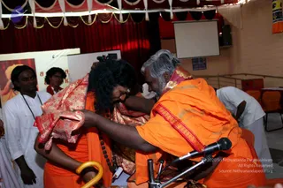 A photograph of a ceremonial moment in an indoor hall with red and white draped fabric overhead and a microphone on a stand in the foreground. A long-haired figure in saffron-orange robes with a red and gold shawl and a dark rudraksha mala bows forward while an older man with swept-back grey hair and a white beard, also in orange robes with a red-bordered sash, presses his hands to the bowed figure's head. The bowed figure holds a curved yellow cane. Two men in white kurtas stand at left, and another person in white bends over chairs at right. A projection screen hangs on the back wall.
