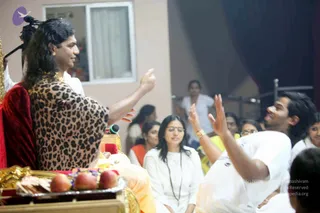 A photograph of a satsang hall where a man with long dark hair, wearing a leopard-print shawl over a red cloth and gold chain, sits on a golden throne and gestures with raised fingers toward a seated audience. Beside him is a silver plate with red-and-green fruit offerings. In the foreground, a man in white clothing raises his hand while speaking, and several women in white and yellow sit listening. A microphone stand is at the upper left and a dark curtain hangs behind. The seated figure shows distinctive long hair, ornate gold necklace and a shawl, but the appearance is not clear enough to confirm identity, so he is described only by role as the seated speaker on the throne.