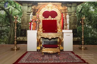 An empty ornate gilded throne with a red velvet cushioned seat and back stands on a red patterned rug, with a matching gilded footstool in front. Behind it is a large printed backdrop of a banyan-like tree and stone temple pillars with carved figures. Two tall brass oil lamps, one on each side, flank the throne. A small figure in orange robes and a floral garland appears on the backdrop's left side near the throne. No people are physically present in the room.