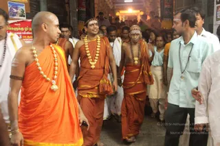 A photograph of a temple corridor crowded with people in a procession-like walk. Three men in the foreground wear saffron-orange robes with gold-coloured borders; the central one has a red-and-gold headband, a long gold bead necklace and a shawl draped over his shoulder, and the man on the right has a shaved head, white sacred marks and a gold pendant. Several men in white dhotis and shirts walk behind them, and a man in a light blue shirt with a rudraksha mala stands to the right. A red banner with Tamil text hangs on the left wall, and a golden-lit shrine entrance glows at the far end.