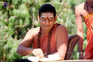 Outdoor photograph of a man seated at a wooden table, eating from a white plate with a metal spoon. He wears an orange-saffron cloth draped over his shoulders, a long rudraksha mala with a ceremonial necklace, and a white vibhuti line with a red kumkum dot on his forehead, plus small gold earrings. Behind him, a second figure in orange garments stands at the edge of the frame beside green leafy foliage.