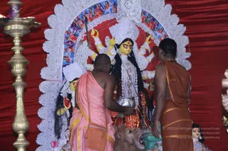 A photograph of a goddess murti in a shrine with a red draped backdrop and a white lace-like arch behind her. The multi-armed deity has yellow skin, a long black braided hair, a white and red adorned costume, and carries weapons in her raised hands, with a lion mount at her base. Two priests stand in front of the murti, both shaved-headed and bare-chested: the left one in a pink dhoti with a yellow border and a crossbody bag, pouring liquid from a silver vessel; the right one in an orange dhoti with a dark orange wrap. A brass oil lamp stands at the left edge. Two people are visible.