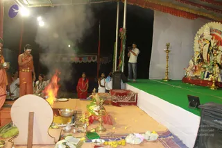 A night-time homa or fire ritual on an open-air stage under a tent canopy. A shaved-headed priest in orange-saffron dhoti and bare upper body stands at left, holding a small vessel, beside a bright sacred fire rising from a pit with offerings. A woman in a white sari with green border and a woman in red sit at the fire. A cameraman in a white shirt and jeans films from the stage. At right, a large decorated deity murti with multiple figures sits on a stage with green cloth, flanked by brass oil lamps. Yellow flowers, fruits, bowls and an open book lie on the mat in front.