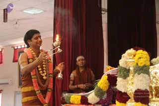 A man in an orange-brown dhoti and shawl, with a rudraksha mala and layered flower garlands in orange, pink and yellow, holds a lit camphor flame in a silver aarti lamp with a handbell in his other hand during a ritual. A second man in saffron robes with a white tilak and a rudraksha mala sits behind him with hands folded in a seated position. Beside them stands a table decorated with white, yellow and red flower garlands and brass vessels, and red curtains hang on the walls. Smoke rises from the lamp, indicating an aarti in progress in an indoor hall.