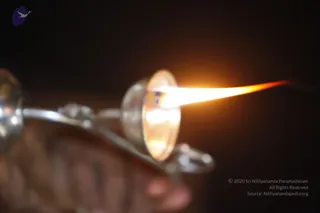 Close-up photograph of a silver aarti lamp with a metal dish and handle, its wick flame blazing in a long orange-yellow tongue against a dark background. The blurred foreground suggests a hand or fabric. No people are visible in the frame.