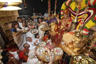 Photograph of a Guru Purnima puja inside a richly decorated shrine room. A man seated on a golden throne with a carved lion base wears an orange-brown turban, a saffron-toned dhoti, a red and yellow floral garland and gold necklaces, and holds his hands in anjali. Devotees around him hold brass plates of offerings and oil lamps; a woman in a blue-trimmed white sari and another in white sari stand at left and centre. Brass bells hang from above and a large deity murti covered in yellow, orange and red flower garlands stands at right. Silver bowls and brass kalasha-style vessels fill a round table in front of the throne, and a cameraman films the ceremony.