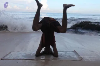 A person performs a headstand on a sandy beach at the water's edge, forearms planted on a flat mat, legs raised straight up into the air. The person wears a dark brown-and-maroon wrap cloth draped over the shoulders and torso. Breaking waves crash behind onto dark rocks under a pale blue sky over the sea.