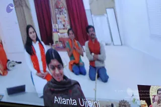 A photograph of a video-call screen showing a satsang link. In the foreground a smiling woman with a red bindi and dark shawl is captioned "Atlanta Ujjaini". Behind her three people in a white-walled room with red curtains kneel and stand with palms pressed together in namaste; a woman in a white top with an orange-red scarf stands at left, a kneeling person in a green and orange top at centre, and a man in a grey sweater with a red-orange scarf at right. On the wall behind them is a small altar with a framed deity image and oil lamps. A smaller inset at lower right shows another screen. The image is a photo of a screen, not a direct scene.