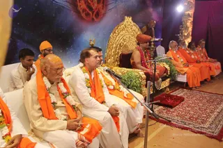 A photograph of a seated assembly on a stage backed by a blue-purple cosmic backdrop with a red glowing sun-like motif. In the centre, a man in white kurta with a saffron shawl and white-and-orange garlands sits before a microphone stand, and an elderly bald man with a yellow-orange tilak on his forehead, white dhoti and white garland sits at left. Behind them, a large ornate golden throne stands empty with a golden peacock finial, flanked by a red-saffron robed figure with a red-orange turban and red-pink garland seated at the throne. Several saffron-robed sadhus with white garlands sit in a row at right, and a figure stands behind the throne near a brass oil lamp. A red patterned carpet lies on the wooden floor.