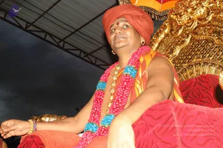 A man sits on an ornate golden throne under a tent canopy at night, wearing a rust-orange turban with a small red tilak on his forehead. He wears a pink and blue flower garland around his neck, a golden necklace, gold bangles, and an orange-and-yellow shawl over his bare upper body. He is seated on a red patterned cushion, with one hand resting on his knee and his gaze lifted upward. Swamiji (probable) is seated as the central figure, with cues including the saffron turban, ornate clothing and garland; the throne suggests a seated honour. The setting is an outdoor canopy with a striped awning and a red-and-yellow umbrella edge at the top.