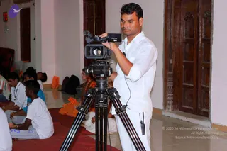 A cameraman in a white shirt stands at a tripod-mounted Sony video camera, filming a seated group in a hall. He is looking through the viewfinder with his hand on the camera body and a small wired microphone clipped to his shirt. Behind him, devotees sit on red mats; a woman in orange robes sits near the centre, and others wear white and grey clothing. Wooden carved doors and a red-lit clock are visible on the wall, and a purple bird logo appears in the top-left corner.