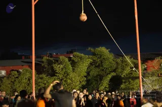 Night outdoor scene of a Janmashtami dahi handi-style celebration: a clay pot hangs from a rope strung between two tall orange-red poles above a crowd. A large crowd of devotees, many in dark t-shirts and some in orange or saffron clothing, gathers below, looking up with raised hands and phones. A person in a dark shirt at the lower left raises a hand toward the pot. Dark trees and red-roofed buildings form the background under a night sky.