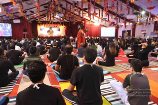 A large tented yoga session seen from behind rows of seated participants in black t-shirts and white-and-black clothes on striped and coloured mats. At the front stands a long-bearded man in a flowing saffron-orange robe, facing the crowd, beneath a stage with a golden deity murti and a backdrop. Two projection screens flank the stage, and the tent ceiling is hung with colourful lanterns and oil lamps. Many participants sit cross-legged in meditation while one woman on the right raises her arms overhead.
