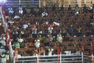 A photograph of a large outdoor evening ritual on tiered stone steps, with many participants seated in rows before rows of black stone shivalingas, each with a small brass oil lamp or kalasha at its base. Lit oil lamps glow across the steps. Many men are bare-chested or wear white dhotis, and women wear white saris, orange, green, and red sarees. Participants sit in meditative posture with hands folded in anjali or pressed together in prayer. Red flags on poles line the left and bottom edges, and a metal chain-link barrier runs along the front. The setting appears to be a large group abhishekam or puja. No single figure is clearly identifiable as a person from appearance.