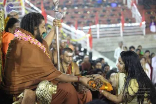 A seated man with long dark hair, a full beard and a swept-back style wears a rust-brown shawl over an orange-brown garment and a pink-and-gold floral garland. He holds a tall silver staff with one hand and extends his other hand to receive a bundle of rudraksha beads tied with an orange cloth from a woman in a cream and gold sari with long dark hair. A gold lion-head armrest is in the foreground. Behind them, saffron-clad attendants and seated devotees fill the stands of a large indoor hall with red flags and white railings. Swamiji (probable) is identified by long swept-back hair, rust-orange shawl and ornate garland cues, seated in the role of the central figure receiving offerings.
