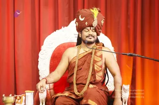 A man seated on an ornate white and red throne against a red drape, eyes closed in meditation. He wears a rust-orange turban with a crescent moon and a yellow feather ornament, a rust-orange shawl over bare shoulders, and a heavy gold necklace. A microphone stand is at his side, and a brass vessel and a steel flask sit on a small table to the left. Swamiji (probable): the turban, ornate gold ornaments and gold necklace are the cues, and the seated figure is a speaker at a satsang.