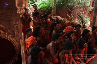 A photograph of a group of devotees seated closely together in an outdoor courtyard with red walls and greenery. Most wear saffron-orange robes and rudraksha malas, many with orange turbans or head cloths, and several sit with palms pressed together in anjali mudra. A young man in the centre-right wears a long-sleeved orange garment with a rudraksha mala and bracelets, hands folded. Behind them are stone and painted tiger figures, and at left an ornately carved wooden or metal pillar with a hanging lamp is visible. The scene suggests devotional gathering or satsang.