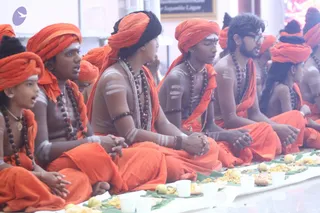 A seated row of young men and boys in saffron-orange dhotis and matching orange turbans sits cross-legged before a long white table set with banana leaves. The leaves hold rice dishes, sweets and fruit, with white cups in front. Several participants wear rudraksha malas and white ash marks on their chests and arms; some have glasses. Their mouths are open as if chanting or speaking.