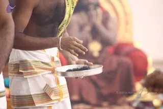 A photograph of a priest's torso and arms during a ritual. He is bare-chested, wearing a white dhoti with multicolored striped borders and a yellow cloth draped over one shoulder, and wears a silver bangle. He holds a round silver plate filled with dark granular material. Behind him, a blurred deity murti sits on a red-draped seat with gold ornaments and a golden halo, with garlands visible. Soft golden light fills the background.