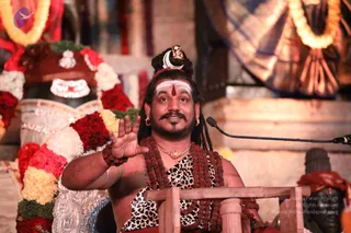 A bearded man with long dreadlocks sits behind a low wooden podium with a microphone on a stand, raising one hand in a gesture while speaking. He wears a crescent moon and small figure ornament in his hair, white and red forehead markings, multiple rudraksha malas, a gold chain, and a leopard-print garment. Behind him a large shivalinga-style form is decorated with red and white flower garlands and gold-trimmed fabric, with a red cloth draped over a brass object at right. The setting is a temple-like hall with warm lighting.