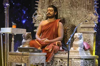 A man in a rust-orange robe with gold trim sits cross-legged in meditation on an ornate gold throne carved with lions and floral motifs, eyes closed and hands resting in his lap. He wears a red tilak on his forehead, a gold necklace and a rudraksha-style bead necklace. A leopard-print cushion sits on the throne seat beside silver lion sculptures. A microphone stands at his right, and a small Yamaha DTX device and an orange cup sit on a silver side table at left against a starry blue backdrop. a person is shown in meditation on his throne during a session.