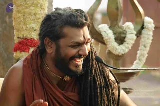 A smiling man with long dreadlocks falling past his shoulders and a red tilak on his forehead speaks into a microphone. He wears a rust-red shawl draped over bare shoulders and a gold chain with a pendant. Behind him, a brass trident is wreathed in white flower garlands, with a red flower garland to the left. The setting appears to be an outdoor satsang. Visible person appearance matches the shawl, dreadlock and trident cues of the page's a person claim, so the identity is probable.