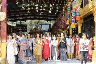 A large group of women and some men stand with palms pressed together in namaste under a open-sided wooden pavilion decorated with hanging colourful tasselled fabric banners and green leaf strands. Among them, a woman in a red kurta with a gold dupatta, a woman in a pink and blue printed salwar, and a woman in a white kurta and grey wrap stand near the front, with golden-yellow carved pillars and a golden shrine structure behind them. Two large screens are mounted on the pavilion walls, and a sign reads Nithyananda Dhyanapeetham Temple area. Many people appear devoted and attentive.