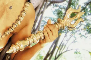 Close photograph of a bare-chested figure holding a gilded trident (trishula) with an ornate golden handle, a hand gripping the shaft. A large beaded rudraksha mala with dark spacers crosses the chest. Blurred green foliage and tree branches fill the background.