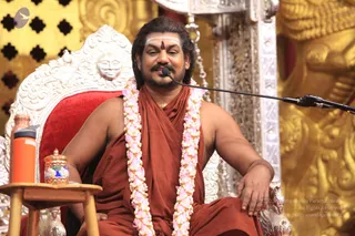 A seated man in saffron-orange robes sits on an ornate silver-and-white throne with a red velvet cushion, speaking into a microphone on a boom stand. He wears a pink and white flower garland, a white tilak with a red dot on his forehead, and a thin gold necklace. His right arm rests on the armrest. Beside him is a small wooden side table holding an orange flask and a decorated tin container. Behind him are gold carved decorations and a gold lattice panel.