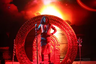 A figure with long dark hair stands on an ornate golden-red throne framed by a large circular halo-like arch, against a glowing orange sunrise backdrop over water. The figure wears a white sleeveless garment, a blue patterned cloth at the waist, a long rudraksha-style mala and beaded necklaces, and bangles on the wrists, while holding a tall staff. A trident stands to the right and a small staff with ornaments to the left. The scene is lit in red and orange light on a stage set, with no other people clearly visible apart from a partial figure at the far left edge. a person appears as the central figure, identified by appearance cues of long swept hair, rudraksha mala and ceremonial adornments.
