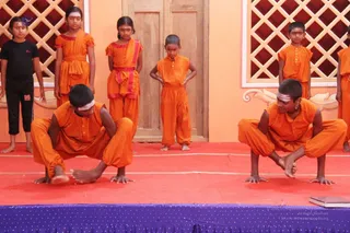 A photograph of a stage performance by children in matching saffron-orange kurta and pant outfits. Two boys in the foreground crouch low on bare feet with hands on the red stage floor, performing a balancing yoga-style posture. Behind them, five children stand in a row, some with hands on hips, against an orange wall with a decorative wooden lattice panel and a wooden door. A plain dark shirt is worn by one girl at left, and a few have tilak on their foreheads. A purple-edged stage apron runs along the bottom.