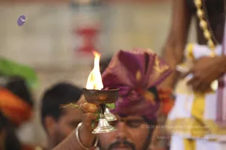 Close view of a priest holding a brass oil lamp with a bright flame, the lamp held up by a raised hand with a silver bangle, during a ritual in a temple setting. He wears a magenta and gold turban and a white garment with gold borders, and a gold necklace hangs at the upper right. Another man with a moustache and orange head cloth stands behind, eyes lowered, and a blurred red and cream structure appears in the background. Offering lamp ritual such as aarti.