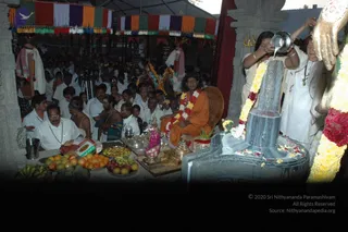A photograph of a temple abhishekam at an outdoor shrine under a canopy hung with multicoloured fringed cloth. A tall Shiva lingam stands at right, being bathed as a woman in white pours liquid from a metal vessel over it, while a flower garland hangs from it. A seated figure in saffron-orange robes with a red-and-white flower garland sits behind a table laden with fruit, bananas, brass vessels and an offering plate, with a person candidate showing long dark hair and saffron clothing. Devotees in white dhotis and shirts crowd the background, and a tripod-mounted camera stands near the group. A trident hangs on the pillar at upper right, and a large metal deity hand is visible at the edge.