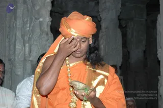 A photograph inside a stone-pillared temple hall with carved pillars behind. A man in an orange turban and orange-and-gold garment, with long dark hair and a heavy gold necklace of beads, presses his right hand to his forehead while holding a leaf cup with greenery in his left hand. Partial figures stand at the edges of the frame, including a man in white at left and an older man in white at right.
