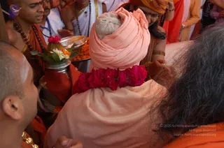 A photograph taken from behind of a seated figure wearing a pale peach turban wrapped around a burlap-covered object, with a large red rose garland draped over the shoulders of the peach shawl. Around them, several people in orange and white clothing crowd in; a man on the left with a shaved head, orange garment and rudraksha beads holds a silver plate of marigolds and a kalasha-like vessel topped with flowers, and another man in white with a rudraksha mala stands behind. A woman in an orange and gold sari with gold bangles stands at right. In the foreground, a shaven-headed man at left wears a gold necklace. The scene suggests a ritual honouring or darshan setting.