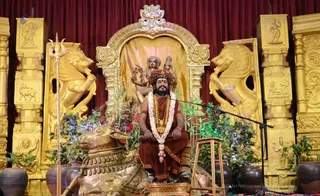 A photograph of a man seated on an ornate gold throne in front of a deep red curtain. He wears rust-orange robes, a saffron head wrap with a tilak mark, a rudraksha mala, a long gold ceremonial necklace, and a white and pink flower garland. He sits behind a gilded Nandi bull statue and a trident leans against the left side. Gold lion figures flank the throne, and green plants and dark pots surround the stage. A microphone stands at right. A gold-colored deity murti with multiple arms rises above the throne. Appearance cues are consistent with a person, seated as a speaker during a satsang talk.