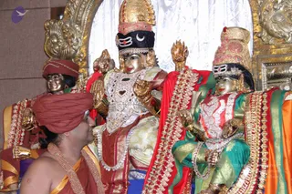 Photograph of a golden deity murti pair seated on an ornate throne, with a central figure wearing a crown with crescent moon and tilak, heavily jeweled white and gold garments, and a red shawl, and a second figure in red and green silk with a tall jeweled crown. A man in the lower left wears a red-orange turban and saffron-orange robe with a beaded mala, seen from behind-side, facing the murtis. Background shows gold ornamental carvings and a white silk backdrop.