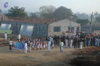A morning procession moves along an unpaved dirt road in front of unfinished concrete-block buildings with a partly built roof frame. Many men in white dhotis and white shirts walk with a line of drummers and dancers wearing white dhotis, red sashes and white caps. Several colourful umbrellas with fringed edges are held above a palanquin area in the middle. A large blue banner with Tamil lettering and a portrait of a saffron-robed figure is carried at the left. Hazy trees and palms fill the background.