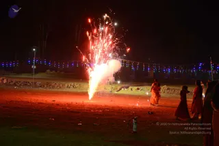 Night photograph of a fireworks display on an open dirt ground at an ashram-style grounds. A large fountain of sparks and white smoke erupts from a firework on the left, with glowing red and orange trails. Several women in saffron and red saris stand and walk on the right, watching the display. Strings of multicoloured fairy lights stretch across the background, and a red fire extinguisher sits on the ground at the lower right. The scene suggests a Deepavali celebration with fireworks.