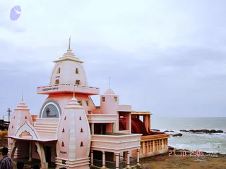 A photograph of a pink and white multi-tiered building with a tall domed tower and spire, set beside a grey sea with rocks along the shoreline under an overcast sky. A red-tiled roof section and small bell-like ornaments appear on the lower pillars. Two partially visible people stand at the lower left edge, one in a blue-and-white checked shirt. The building bears Tamil lettering on its upper facade and a circular wheel emblem above the entrance arch.